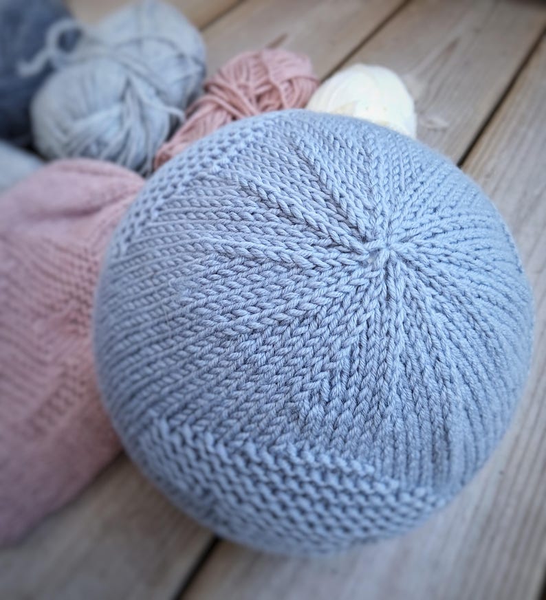 K&ouml;nnte beinhalten: Ein hellblaues, rundes Strickkissen mit strukturierter Oberfl&auml;che. Im Hintergrund befinden sich mehrere Wollkn&auml;uel in Grau-, Rosa- und Wei&szlig;t&ouml;nen. Das Kissen ist aus weichem Garn gefertigt und wahrscheinlich f&uuml;r die Inneneinrichtung gedacht.