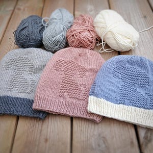 K&ouml;nnte beinhalten: Drei gestrickte Baby-M&uuml;tzen in Hellblau, Rosa und Grau, jeweils mit einem Dinosaurier-Design. Die M&uuml;tzen werden mit Wollkn&auml;ueln in passenden Farben pr&auml;sentiert. Die M&uuml;tzen haben einen umgeschlagenen Saum und sind aus weichem Garn.