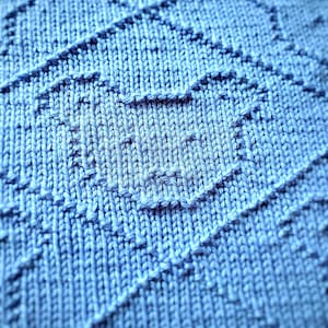 May include: Close-up of a knitted blue blanket with a textured pattern. The blanket features a repeating geometric design with diagonal lines and a heart-shaped motif. The soft, light blue color and detailed stitching create a cozy, handmade aesthetic.