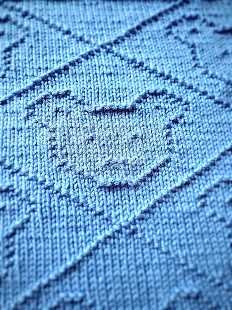 May include: Close-up of a knitted blue blanket with a textured pattern. The blanket features a repeating geometric design with diagonal lines and a heart-shaped motif. The soft, light blue color and detailed stitching create a cozy, handmade aesthetic.