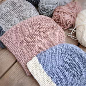 K&ouml;nnte beinhalten: Nahaufnahme von mehreren gestrickten M&uuml;tzen in verschiedenen Farben, darunter Grau, Rosa und Blau, mit einem strukturierten Muster. Die M&uuml;tzen werden zusammen mit Wollkn&auml;ueln in passenden Farben pr&auml;sentiert, was auf ein handgemachtes Bastelprojekt hindeutet. Die M&uuml;tzen haben einen Umschlag.