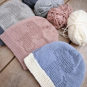 K&ouml;nnte beinhalten: Eine Sammlung handgestrickter M&uuml;tzen in verschiedenen Farben, darunter Grau, Rosa und Blau, mit einem strukturierten Muster. Mehrere Wollkn&auml;uel in passenden Farben sind daneben angeordnet, was auf ein Bastelthema hindeutet. Die M&uuml;tzen werden auf einer Holzoberfl&auml;che pr&auml;sentiert.
