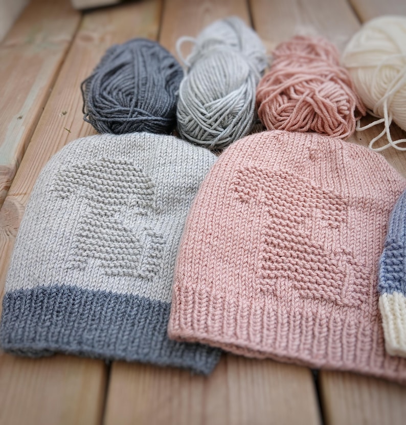 K&ouml;nnte beinhalten: Handgestrickte M&uuml;tzen in verschiedenen Farben, darunter Grau, Rosa und Blau, mit einem strukturierten Design. Die M&uuml;tzen werden zusammen mit Wollkn&auml;ueln in passenden Farben pr&auml;sentiert, was auf ein Bastel- oder DIY-Thema hindeutet.