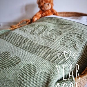 May include: A sage green knitted blanket with the year "2026" knit into the fabric. The blanket has a heart pattern and is draped over a basket. A brown teddy bear sits on top of the blanket.