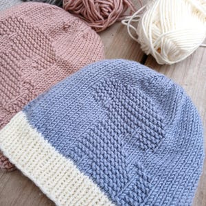 K&ouml;nnte beinhalten: Nahaufnahme von handgestrickten M&uuml;tzen in Blau-, Rosa- und Graut&ouml;nen, mit wei&szlig;en Garnkn&auml;ueln im Hintergrund. Die M&uuml;tzen haben ein strukturiertes Strickmuster und einen wei&szlig;en Rand. Das Bild zeigt die Handwerkskunst des Strickens.