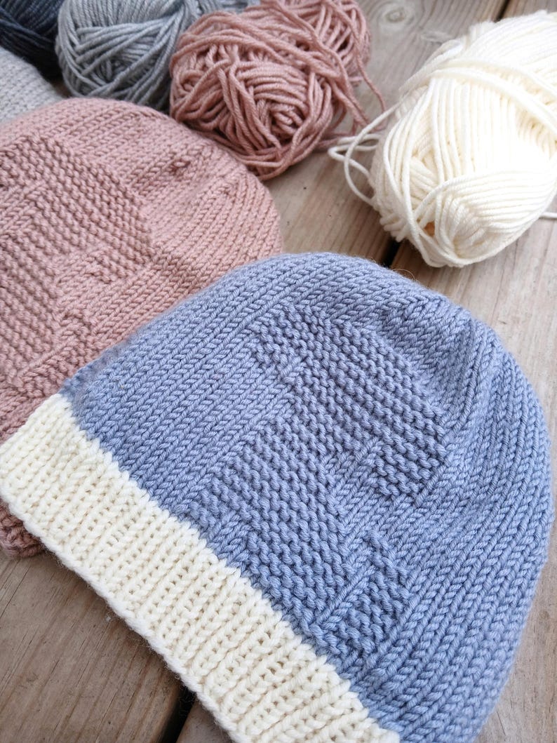 K&ouml;nnte beinhalten: Nahaufnahme von handgestrickten M&uuml;tzen in Blau-, Rosa- und Graut&ouml;nen, mit wei&szlig;en Garnkn&auml;ueln im Hintergrund. Die M&uuml;tzen haben ein strukturiertes Strickmuster und einen wei&szlig;en Rand. Das Bild zeigt die Handwerkskunst des Strickens.