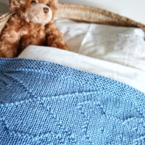 May include: A light blue knitted blanket with a geometric pattern is draped over a bed. A brown teddy bear sits on the pillows behind the blanket. The blanket is made of soft yarn.