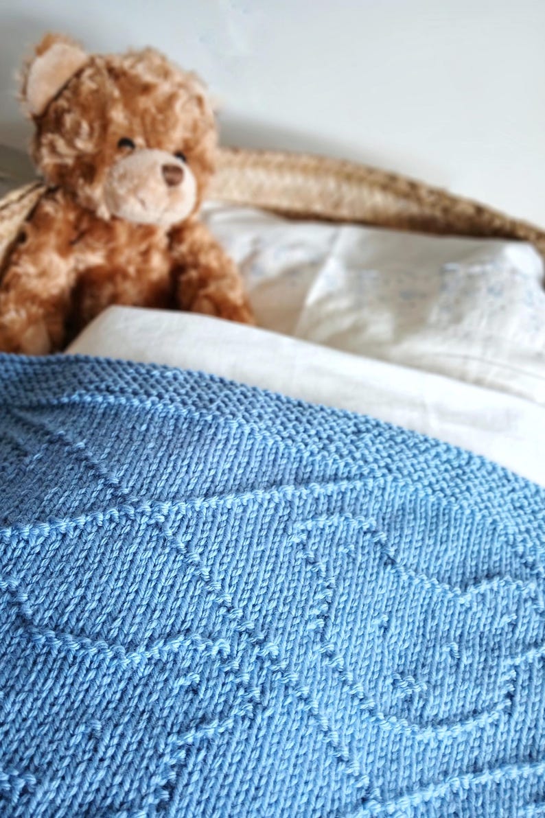 May include: A light blue knitted blanket with a geometric pattern is draped over a bed. A brown teddy bear sits on the pillows behind the blanket. The blanket is made of soft yarn.