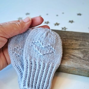 May include: A pair of light gray knit baby mittens featuring a subtle heart-shaped pattern on the back of each hand. The mittens are constructed with a ribbed cuff and a soft, textured yarn, designed to provide warmth for an infant's hands.
