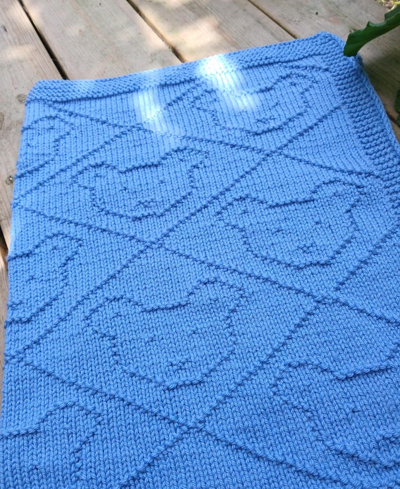 May include: A knitted baby blanket in a light blue color. The blanket features a repeating pattern of bear faces within a diamond grid. The edges are neatly finished with a ribbed border, creating a cozy and handmade feel.