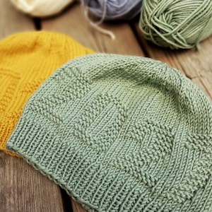 May include: A sage green knitted beanie featuring the word LOVE knitted into the fabric using a textured stitch pattern. The beanie has a ribbed brim and is positioned next to a yellow knitted beanie and several balls of yarn in various colours.