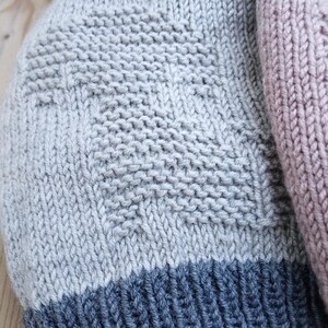 K&ouml;nnte beinhalten: Nahaufnahme von drei gestrickten M&uuml;tzen. Eine ist hellgrau mit dunkelblauem Rand, eine weitere hellrosa und eine dritte grau. Die M&uuml;tzen haben ein strukturiertes Strickmuster.