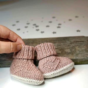 May include: A pair of knitted baby booties in a light pink color with a white trim. The booties have a ribbed texture and are made with a soft yarn.