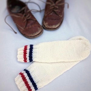 May include: A pair of white knitted leg warmers with red, white, and blue stripes at the top. The leg warmers are folded in half and are lying on a white surface.