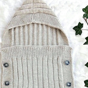 Puede incluir: Un saco de dormir para bebé de punto beige con capucha y botones. El saco de dormir está hecho de un material suave y cómodo, perfecto para mantener a los bebés calientes y cómodos.