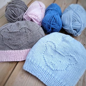May include: A collection of hand-knitted beanies and yarn. The beanies are gray and light blue, each featuring a heart design. The yarn is in shades of gray, pink, blue, and light blue. The items are arranged on a wooden surface.