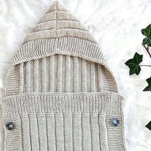 May include: A beige knitted hooded sleep sack featuring a vertical ribbed texture throughout the body and hood. The garment includes four dark, circular buttons positioned symmetrically along the sides to secure the front flap in place.