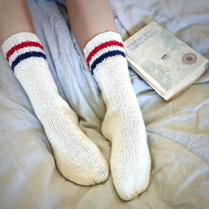 Pode incluir: Um par de meias brancas de malha com listras vermelhas e azuis em torno da parte superior. As meias estão sobre uma colcha branca com um livro em cima.