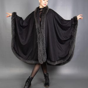 May include: A black cape with grey fur trim around the collar and edges. The cape is worn over a black outfit and black boots. The model is posing with arms outstretched against a grey background.