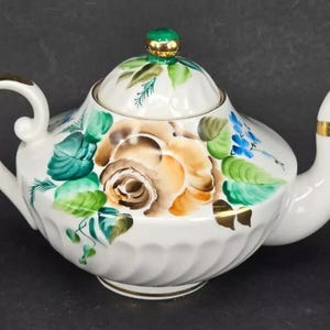 Pode incluir: Bule de porcelana branca com um design floral. A chaleira tem uma tampa verde com uma pega dourada e uma borda dourada em torno do bico. O design floral apresenta flores amarelas, laranja, verdes e azuis.