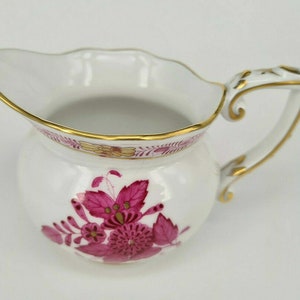 Può includere: Una lattiera in porcellana bianca con corpo rotondo e bordo smerlato. La lattiera presenta un motivo floreale magenta e finiture dorate lungo il bordo e il manico. Il manico ha un design decorativo.