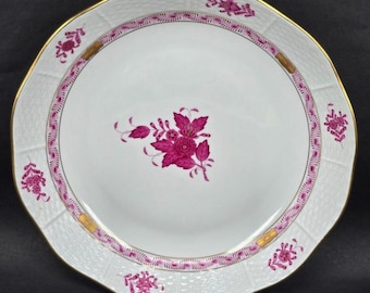May include: A white porcelain dinner plate with a pink floral design and a gold rim. The plate has a scalloped edge and a raised pattern around the rim.