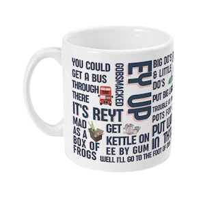 Può includere: Una tazza in ceramica bianca con un design blu e rosa con il testo "You could get a bus through there, it's reyt mad as a box of frogs, gobsmacked, ey up, big do's & little do's, put big lights, trouble for a kettle on, put water in the pots for a well I'll go to the foot of our".