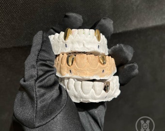 May include: Three sets of gold teeth grills, stacked on top of each other, in a black glove. The grills are made of gold and have a shiny finish.