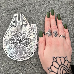 Puede incluir: Una pegatina plateada con un contorno en blanco y negro del  Millennium Falcon. La nave espacial está rodeada por un diseño de mandala. La pegatina está al lado de una mano con esmalte de uñas verde y un tatuaje de un diseño de mandala en la muñeca.