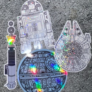 Pode incluir: Quatro adesivos holográficos com personagens de Star Wars. Os adesivos são desenhos em preto e branco com um acabamento arco-íris iridescente. Os personagens são R2-D2, um sabre de luz, a Estrela da Morte e o Millennium Falcon.