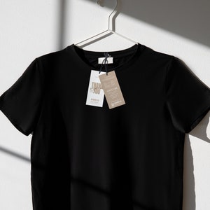 May include: A black crew neck t-shirt hanging on a white hanger against a white wall. Two tags are attached to the shirt. One tag says "THANK YOU" and the other tag says "HOW TO CARE".
