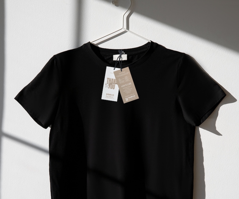 May include: A black crew neck t-shirt hanging on a white hanger against a white wall. Two tags are attached to the shirt. One tag says "THANK YOU" and the other tag says "HOW TO CARE".