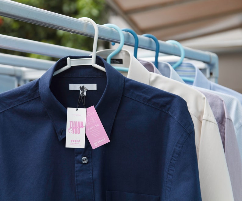 May include: Several button-down shirts hang on a clothesline. The front shirt is navy blue with a white tag that reads "THANK YOU". Other shirts are white, light blue, and striped. The shirts are on white and blue hangers.