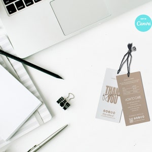 May include: Two clothing tags with black strings, one white and one tan, lie on a white surface near a laptop and stationery. The white tag says "THANK YOU" in gold script. The tan tag includes size and care instructions.