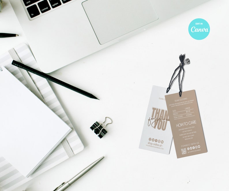 May include: Two clothing tags with black strings, one white and one tan, lie on a white surface near a laptop and stationery. The white tag says "THANK YOU" in gold script. The tan tag includes size and care instructions.