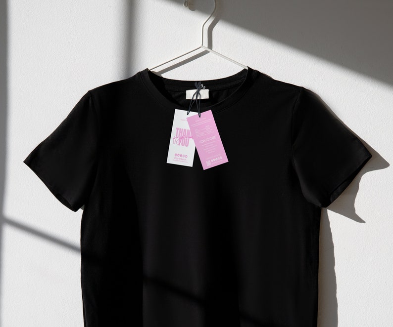 May include: A black crew neck t-shirt hanging on a white hanger against a white wall. Two tags are attached to the shirt. One tag is white with the words "THANK YOU" in pink. The other tag is pink with text.