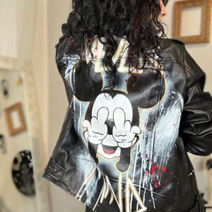 Könnte beinhalten: Schwarze Lederjacke mit einem weißen und schwarzen Mickey-Maus-Design. Mickey Mouse zeigt mit beiden Händen den Mittelfinger. Die Jacke hat goldene und rote Farbspritzer.