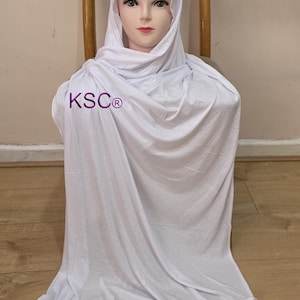 Pode incluir: Um hijab branco e tecido a condizer drapeado sobre a cabeça e ombros de um manequim. O tecido é macio e fluido, com o texto "KSC®" visível na frente. O manequim está sentado numa cadeira de madeira.