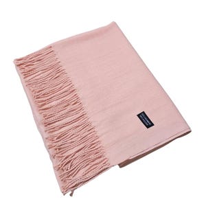 Op de afbeelding: Een lichtroze sjaal met franjes aan één kant. De sjaal is rechthoekig en gemaakt van zacht materiaal. Een klein zwart label met witte tekst is zichtbaar. De sjaal is opgevouwen en plat neergelegd.