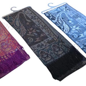 Puede incluir: Tres bufandas rectangulares con estampados de cachemira y flecos en los extremos, presentadas en perchas de plástico. Las bufandas son de tonos morado y naranja, negro y gris, y azul claro y oscuro, cada una con una pequeña etiqueta rectangular.