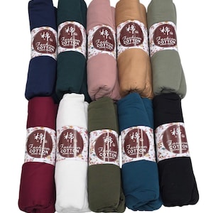 Può includere: Una collezione di sciarpe di cotone arrotolate in vari colori, tra cui blu navy, verde, rosa, beige e oliva. Ogni sciarpa è avvolta con un'etichetta che recita "Fashion Cotton Hand Made."