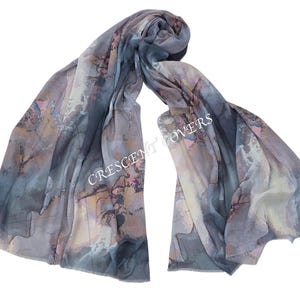 May include: A flowing scarf with a watercolor-like design in shades of blue, gray, and peach. The scarf has a soft, draped appearance and features the text "CRESCENT COVERS" in white. The fabric appears lightweight and suitable for various seasons.
