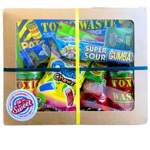 May include: A brown cardboard box filled with various colourful sour candy packages. The box has a clear window to show the candy inside. The candy includes packages of Toxic Waste, Warheads, Super Sour Gumball, and Q4 Chewz.