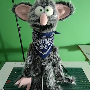 May include: A gray puppet with large, round eyes and a pink nose and ears. The puppet has a blue bandana around its neck and is made of fluffy material. The puppet is sitting on a green cutting mat.