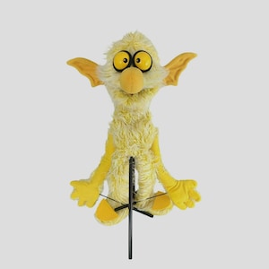 Könnte beinhalten: Eine gelbe Handpuppe mit zotteligem Fell, großen runden Augen mit schwarzen Pupillen und spitzen Ohren. Die Puppe hat eine knollige Nase, glatte gelbe Hände und Füße, die an einer schwarzen Metallstange mit dünnen Drähten befestigt sind.
