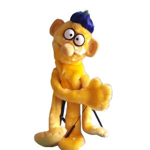 Può includere: Burattino di peluche giallo fatto a mano con un'acconciatura blu. Il burattino ha grandi occhi espressivi, un naso bulboso e grandi orecchie. Ha braccia e gambe lunghe, con corde nere attaccate per la manipolazione. Il burattino è su sfondo bianco.