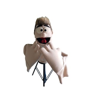 Könnte beinhalten: Eine beige Puppe mit menschenähnlichem Gesicht, braunen Haaren und großen Augen. Die Puppe hat einen weit geöffneten Mund mit roter Zunge und ist mit erhobenen Händen posiert. Die Puppe ist auf einem schwarzen Ständer montiert.