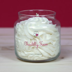 Peut inclure: Une bougie en verre transparent avec une cire blanche en forme de crème fouettée. L'étiquette indique "Chantilly Fraise" et "Fabriqué à Chantilly".
