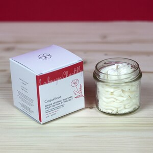 Peut inclure: Une bougie en pot blanc avec une cire blanche ressemblant à de la crème fouettée. La bougie est étiquetée "Coquelicot" et est une bougie parfumée végane. Le pot est posé sur une surface en bois.