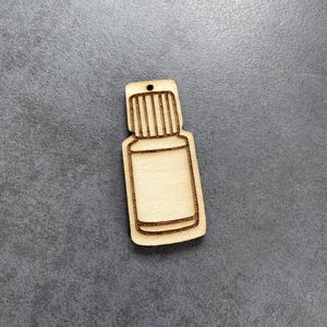 Könnte beinhalten: Ein hölzerner Ausschnitt in Form einer kleinen ätherischen Ölflasche. Die Flasche hat einen rechteckigen Körper und einen Deckel mit vertikalen Linien. Oben befindet sich ein kleines Loch zum Aufhängen. Das Holz hat eine helle, natürliche Farbe mit dunkelbraunen Umrissen.